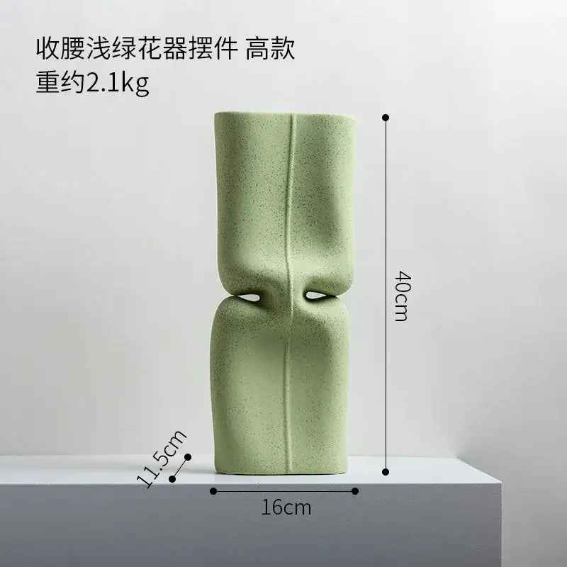 A sleek matte sage green hollow ceramic vase with sculpted hourglass silhouette and subtle vertical ridges for living room decor