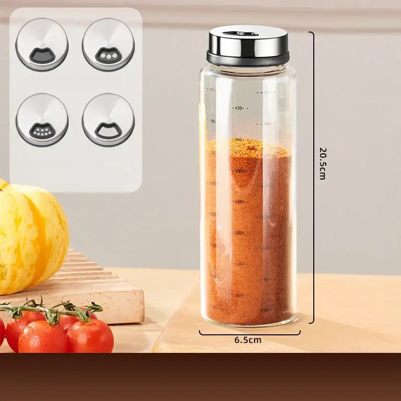 Clear glass seasoning jar with silver metal lid and measurement markings filled with orange-red seasoning in a modern set