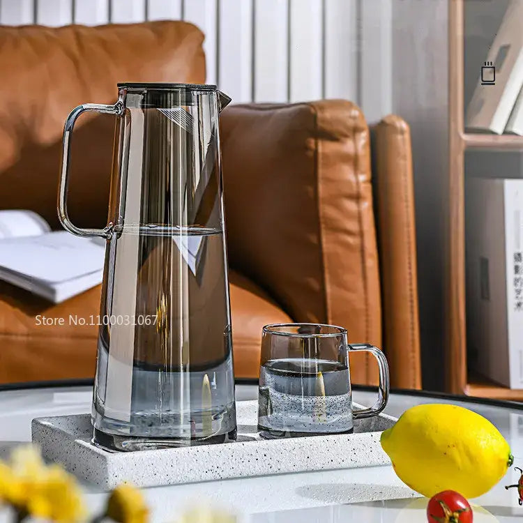 Modern silver glass water pitcher with sleek handle and black lid beside matching glass water mug on speckled tray
