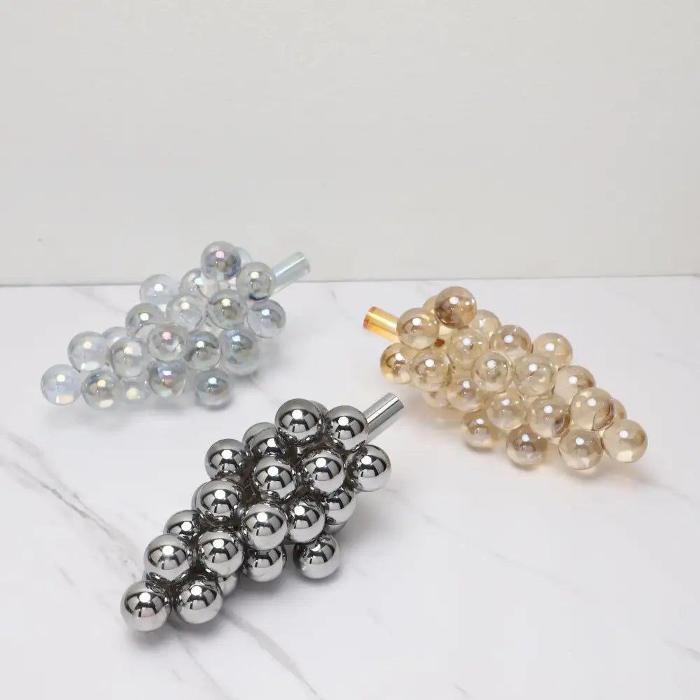 Three metallic crystal grape decorative sculptures in silver gold and chrome finishes on a marble surface