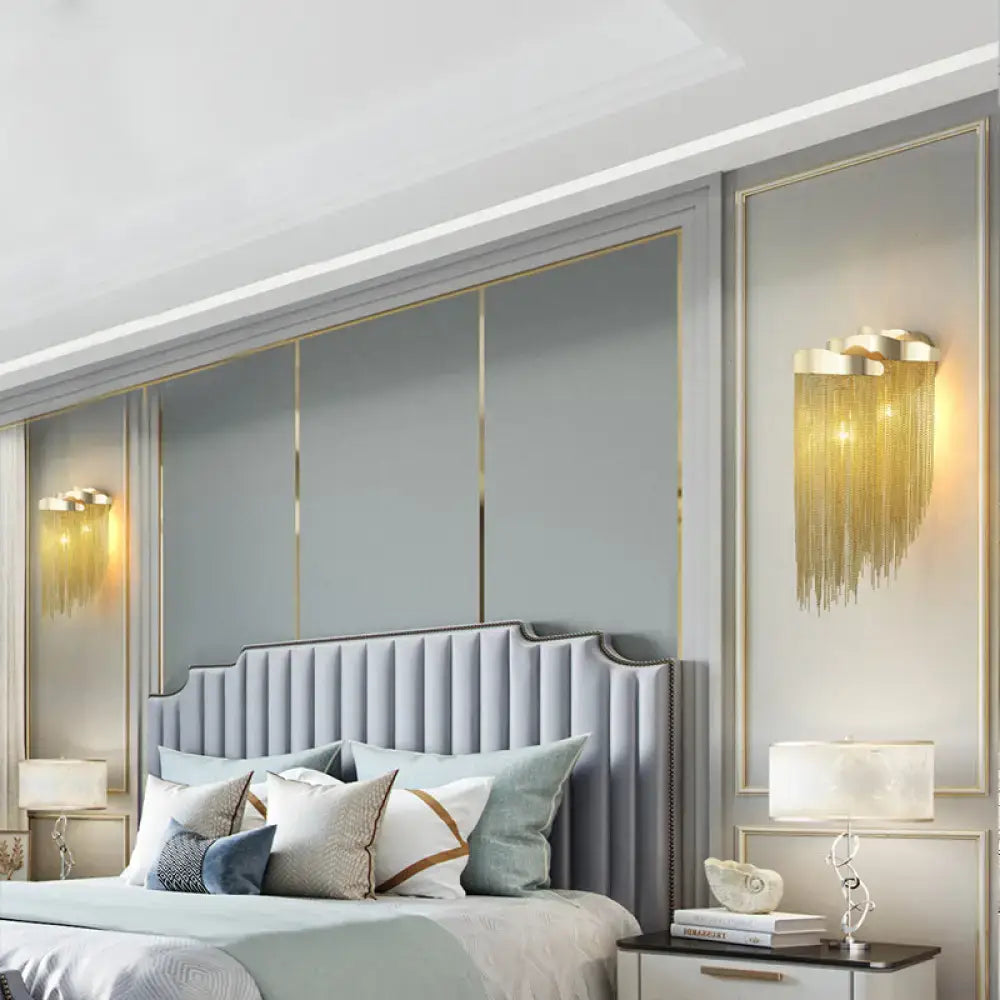 Modern silver and gold tassel design wall lamp with bulb base above a light gray tufted bed with curved headboard and pillows