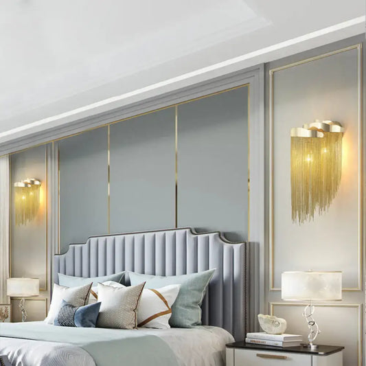 Modern silver and gold tassel design wall lamp with bulb base above a light gray tufted bed with curved headboard and pillows