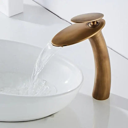 Antique brass waterfall basin faucet with a sleek curved spout and flat circular handle for modern bathroom decor
