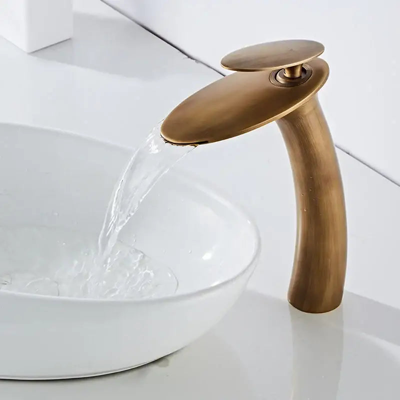 Antique brass waterfall basin faucet with a sleek curved spout and flat circular handle for modern bathroom decor