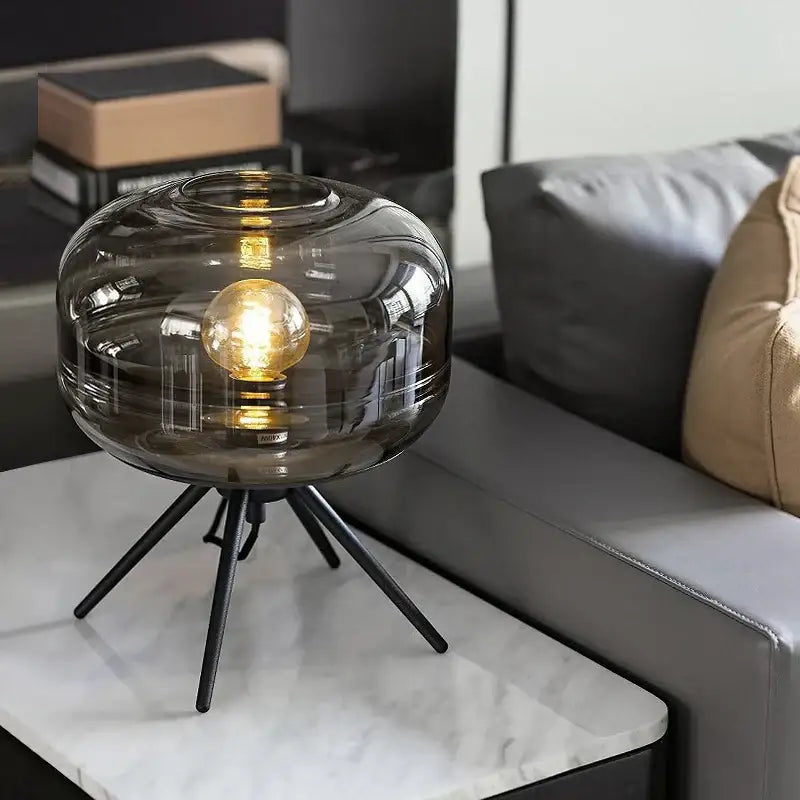 A modern smoked glass desk lamp with tripod base and exposed golden bulb from decorbites glass desk collection