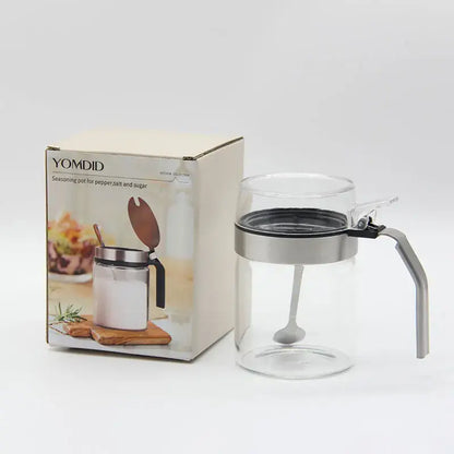 Clear glass seasoning pot with silver metal lid and handle featuring an integrated spoon for kitchen supplies