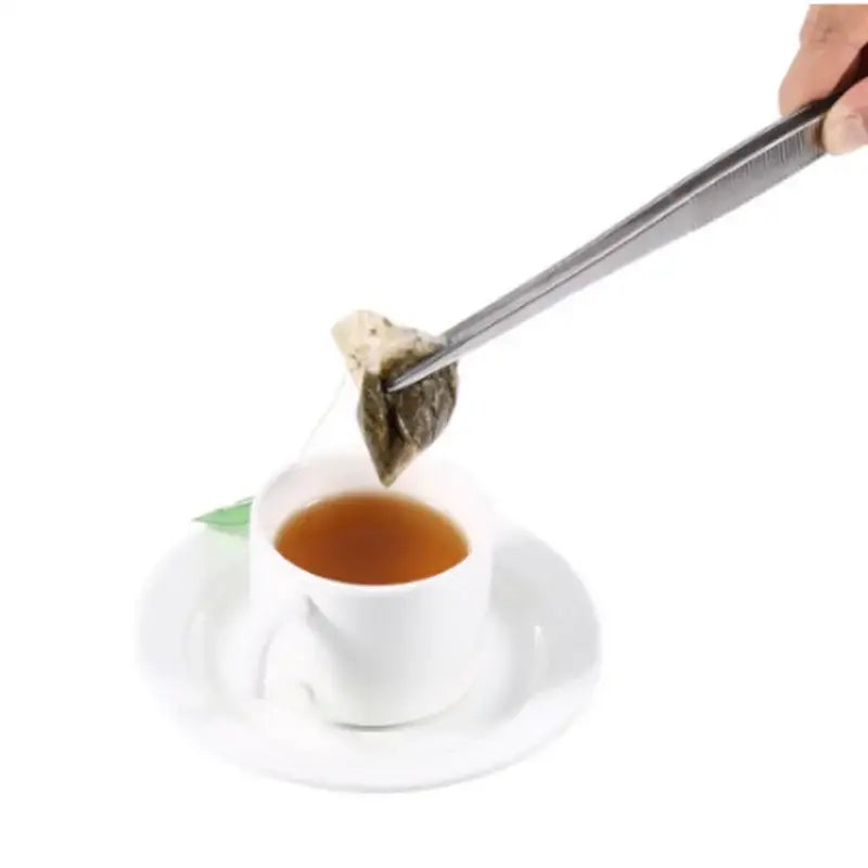 A stainless steel bbq tongs with textured grip holding a dark leaf-shaped teabag over a white ceramic cup