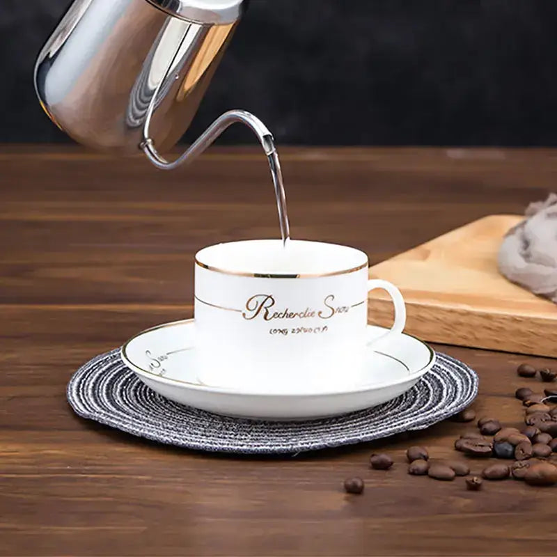 White porcelain coffee cup with gold trim on matching saucer beside decorbites gooseneck stainless steel kettle