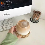 A hand holds a transparent glass coffee mug filled with iced coffee resting on a woven coaster for dining use