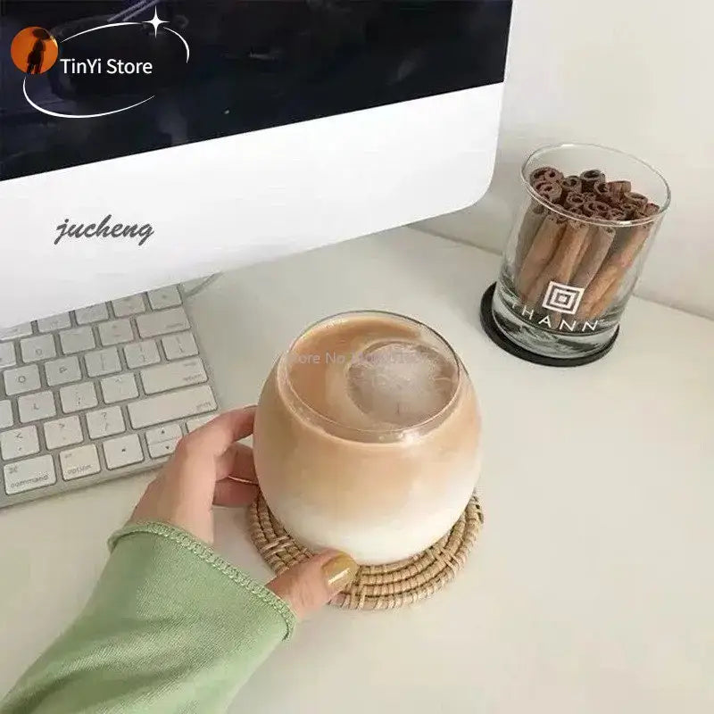 A hand holds a transparent glass coffee mug filled with iced coffee resting on a woven coaster for dining use