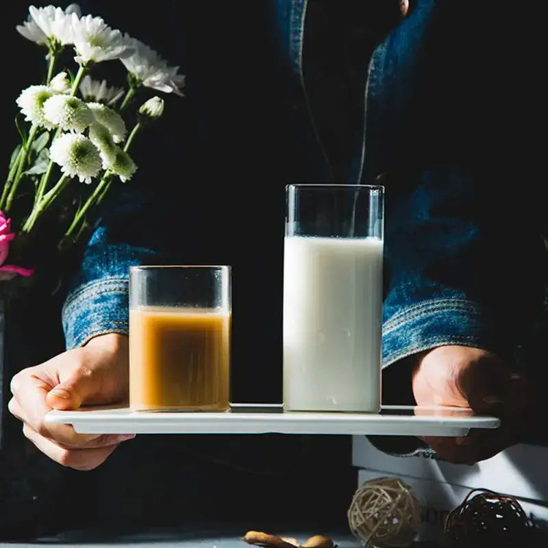 A white ceramic tray holding two transparent glass square cups one filled with creamy beige liquid and the other with milk
