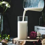 A modern transparent glass square measuring cup pours creamy white liquid into a tall transparent glass cup