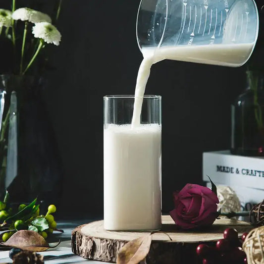 A modern transparent glass square measuring cup pours creamy white liquid into a tall transparent glass cup