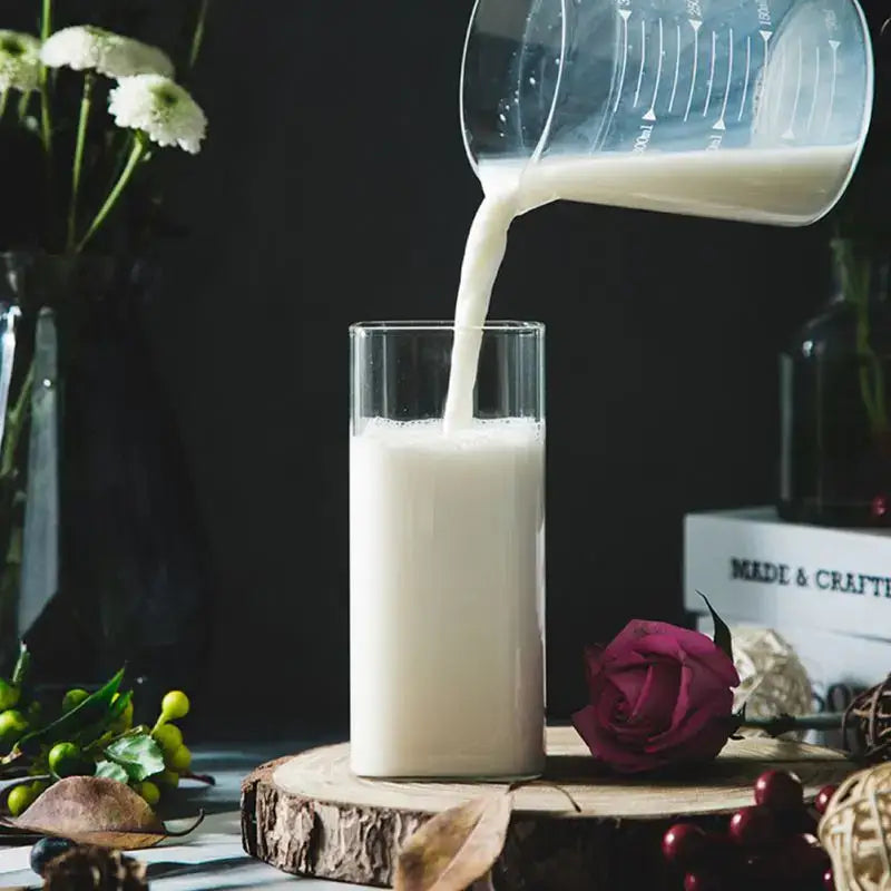 A modern transparent glass square measuring cup pours creamy white liquid into a tall transparent glass cup