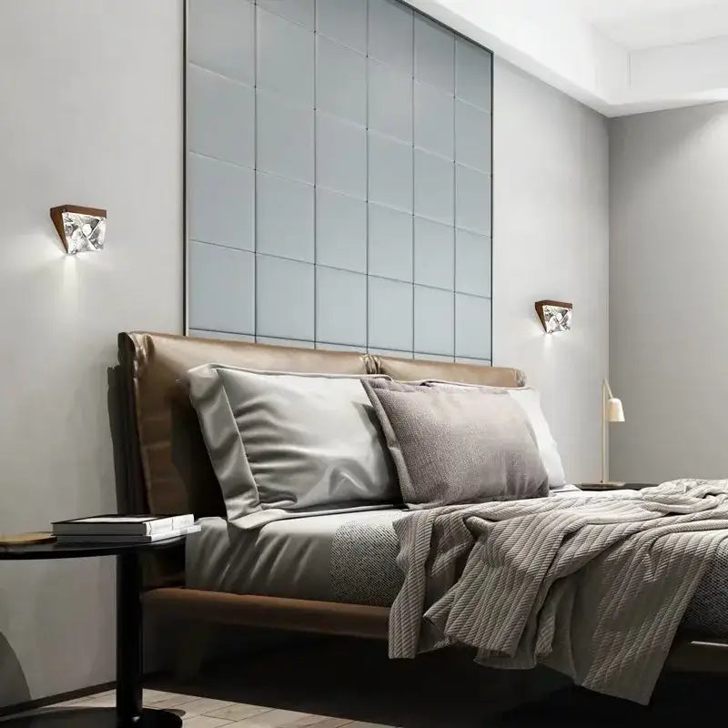 Modern triangular bronze crystal wall sconce installed above a brown leather bed with gray pillows and textured blanket