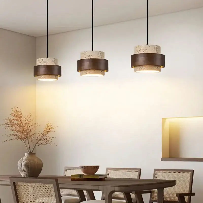 Three modern beige and dark bronze stone pendant lights with speckled cylindrical shades hang above a dining table