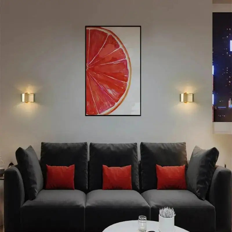 A modern wall lamp in black gold is installed above a sofa with a vibrant red grapefruit watercolor painting in a black frame