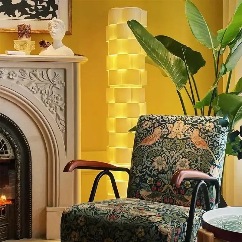 A modern white cylindrical vertical floor lamp next to a dark green plush armchair with floral and bird motifs