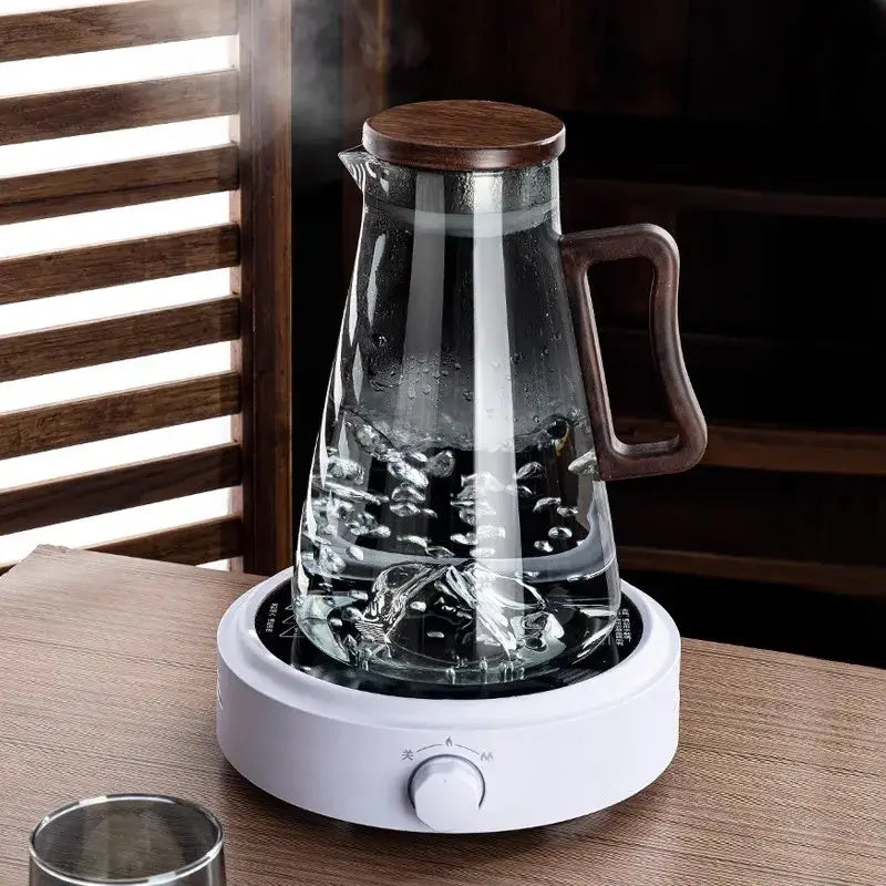 Clear glass cold water jug with modern dark wood handle and lid, sitting on a white electric kettle base