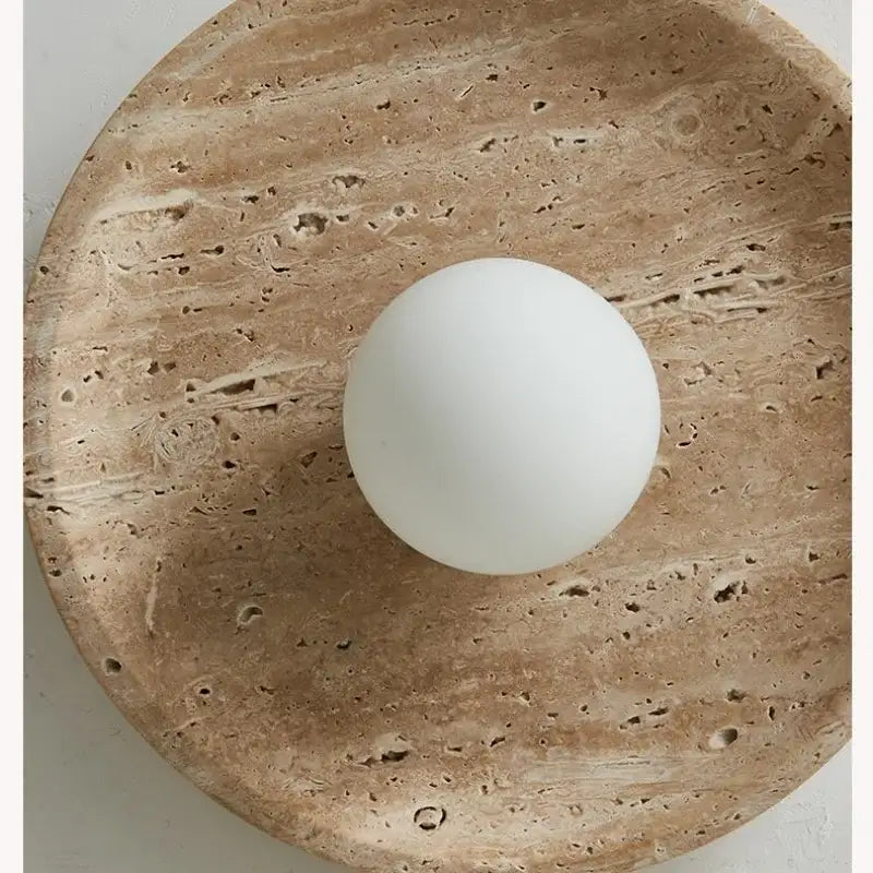 A smooth matte white glass ball led ceiling light resting on a textured beige yellow travertine stone plate decorbites glass ball