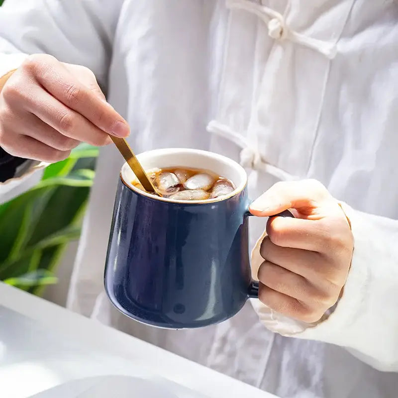 Deep blue ceramic mug with white rim from a navy and emerald ceramic mug set used for stirring iced coffee with gold spoons