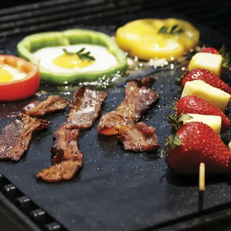 Black non-stick stone bbq grill mat with bacon eggs in bell peppers and fruit skewers cooking on grill mat