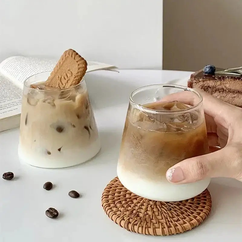 Clear glass iced coffee cup with frothy base and ice cubes held on woven coaster from nordic pink glass cocktail cups set decorbites glass cocktail