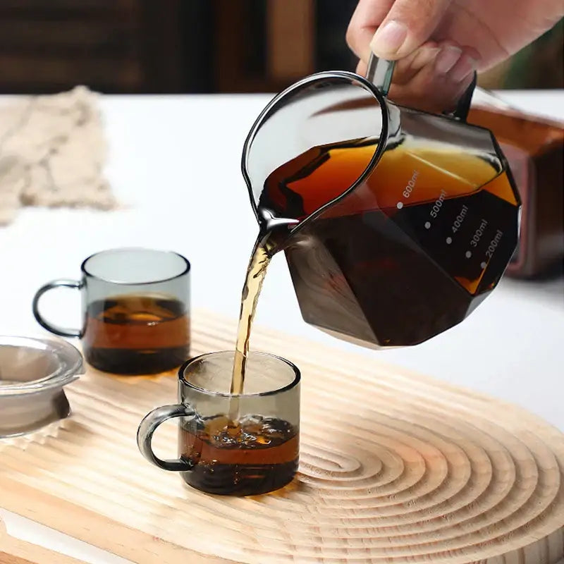A dark amber glass coffee kettle octagonal with a faceted design and measurement markings pouring into matching cups