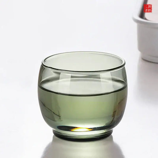A translucent olive green decorbites glass tea cup with rounded base and smooth rim in a kung fu tea cup set