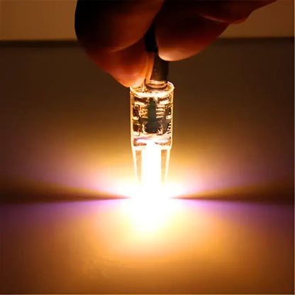 A hand holds a glowing clear glass orange decorbites G4 led bulb 220v cob light with two prongs at the base