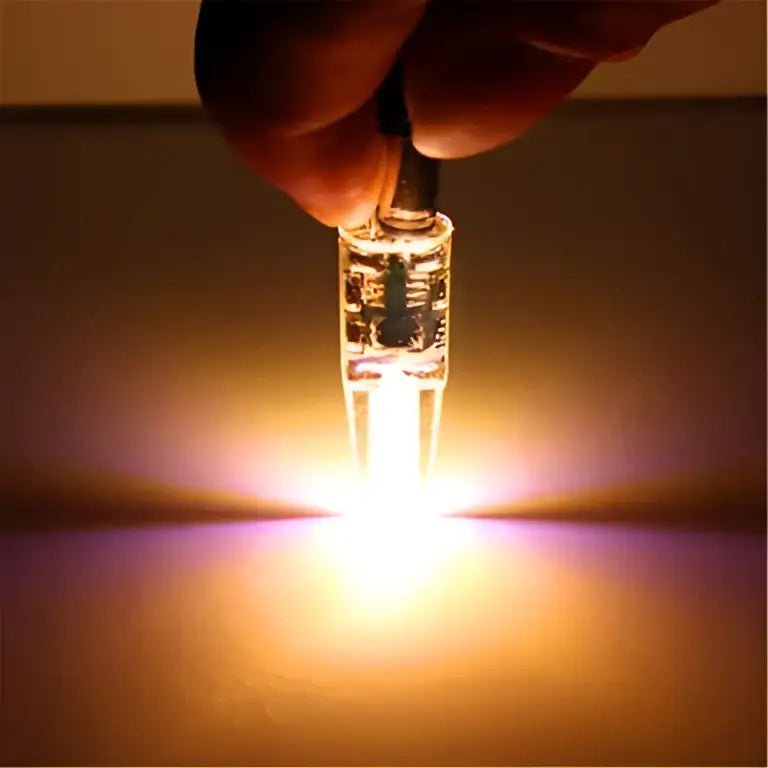 A hand holds a glowing clear glass orange decorbites G4 led bulb 220v cob light with two prongs at the base