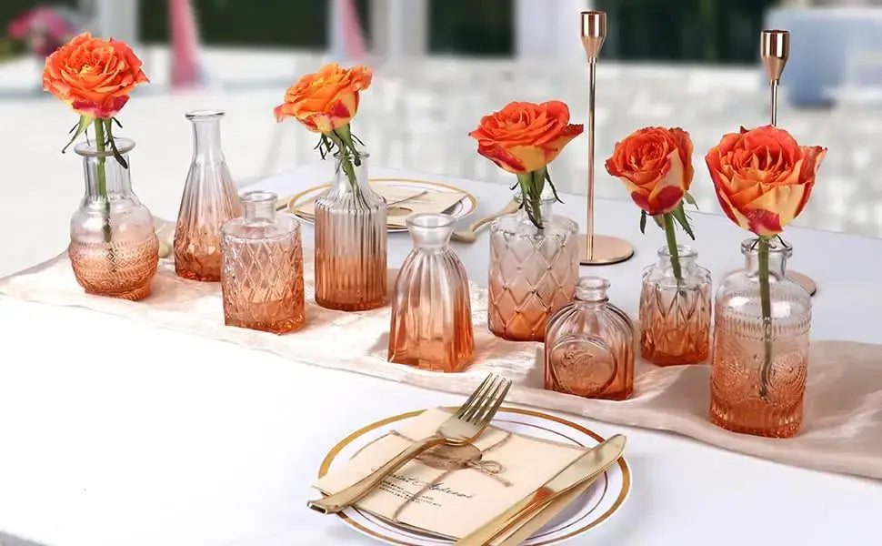 A collection of rose-gold gradient glass bud vases with textured patterns holding single orange roses for wedding centerpieces