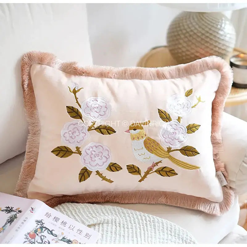 A soft blush pink decorbites bamboo embroidered fringed pillow cover featuring white flowers green leaves and a yellow bird