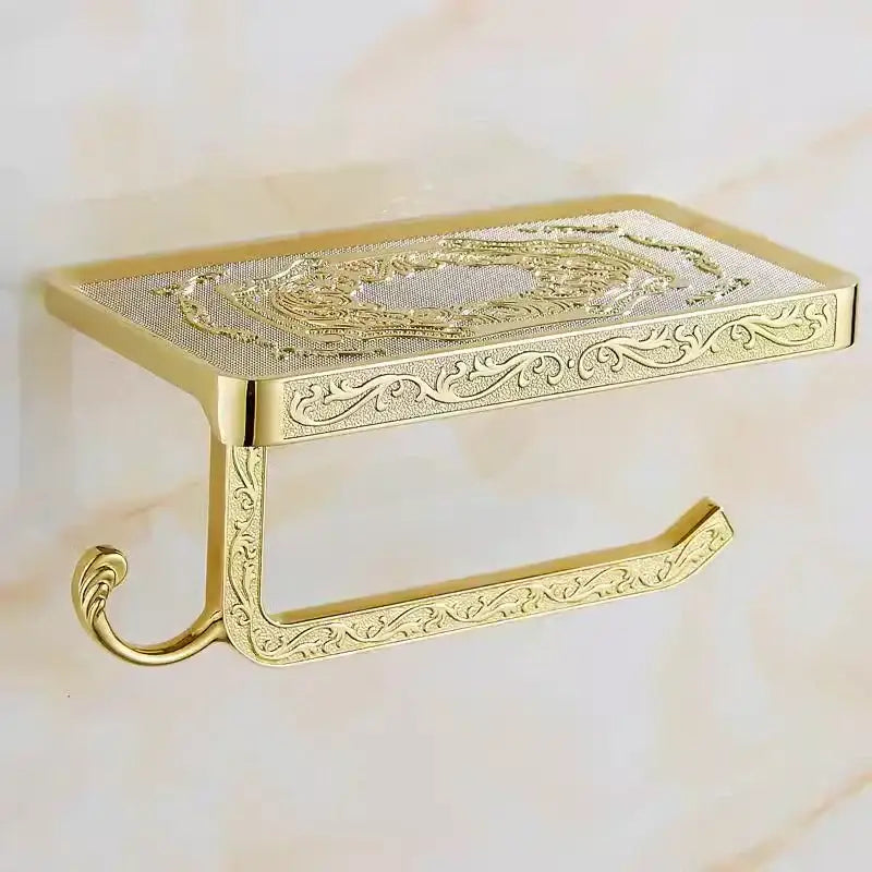 Gold-toned metal toilet paper holder with ornate embossed floral scrollwork and curved support arm for bathroom use