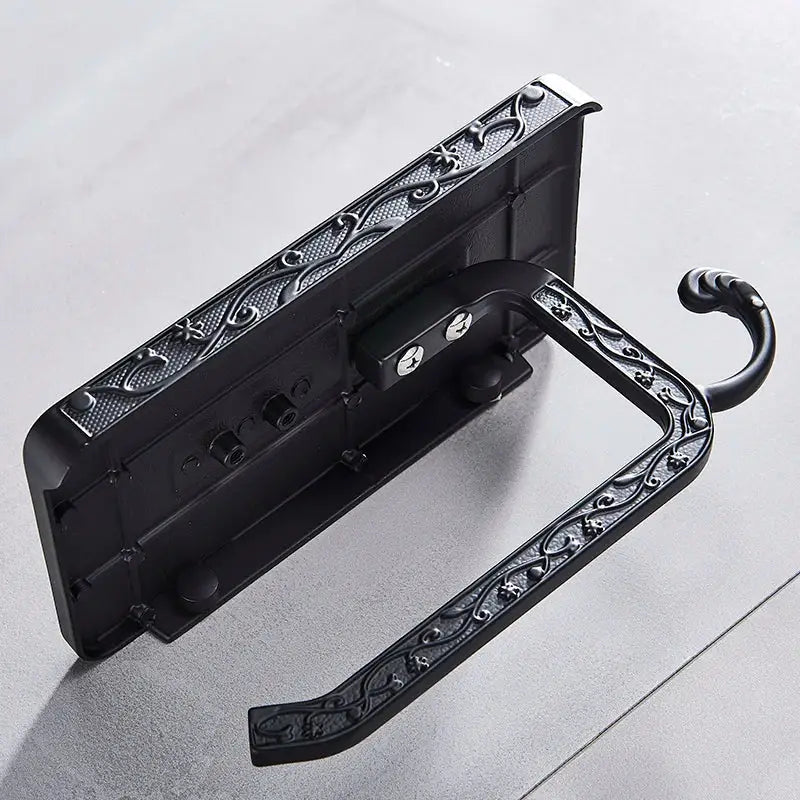 Ornate black metal toilet paper holder featuring intricate scrollwork design with phone shelf for bathroom storage