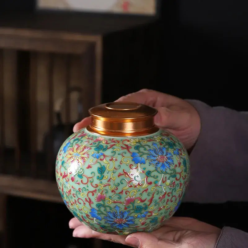 A hand holds a light green ceramic storage jar with ornate blue and pink floral patterns and a gold lid for tea
