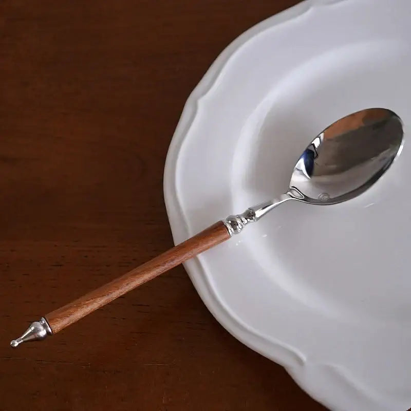 A sleek silver stainless steel spoon with polished wooden handle and ornate accents on a white ceramic plate in a western tableware set