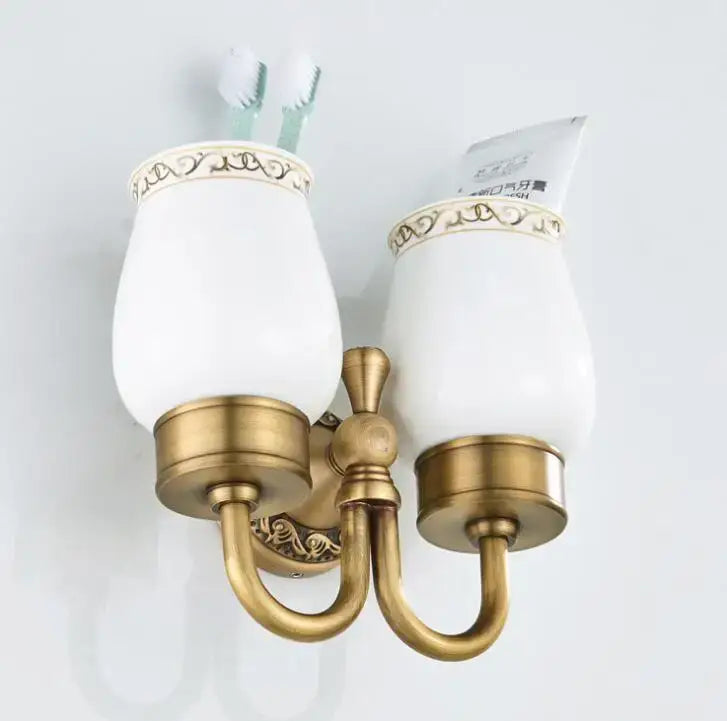 Antique brass-finished wall-mounted double cup holder with white ceramic cups and ornate gold-trimmed rims for toothbrushes