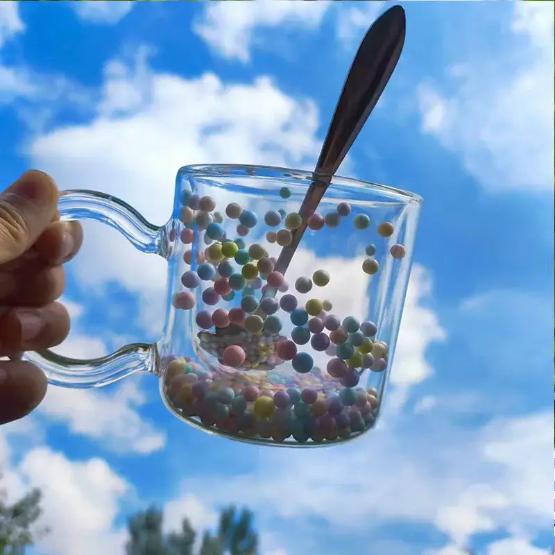 A clear double wall glass cup with pastel heart bubble quicksand spheres and spoon against a bright blue sky