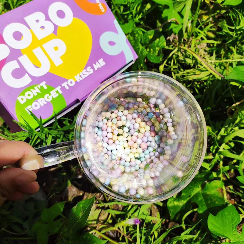 A clear pastel-filled heart bubble quicksand double wall glass cup held by hand against green grass background