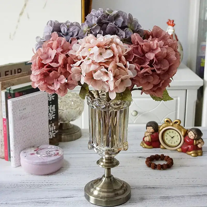 A clear faceted glass vase with metallic base holds pastel pink and lavender decorbites hydrangea branch silk flowers
