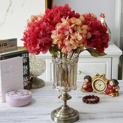 A tall clear glass vase with ornate silver base holding a pastel decorbites hydrangea branch silk flower bouquet