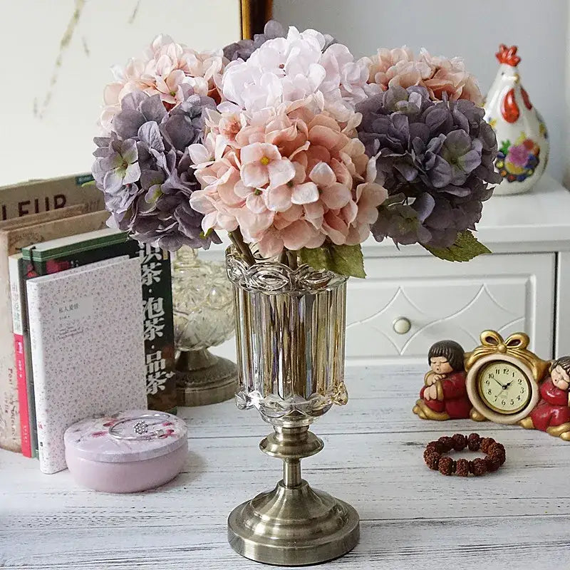 A tall clear glass vase with antique brass base holding a pastel decorbites hydrangea branch silk flower bouquet