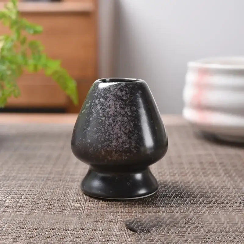 A sleek black ceramic tea ceremony scoop with speckled glaze and rounded base in pastel pink and yellow matcha whisk stand set