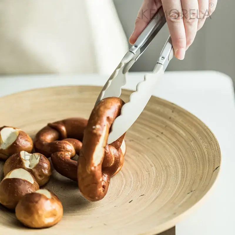 Silver-toned kitchen tongs with textured non-slip grip lifting pretzel as part of pastel silicone kitchen utensils set with ceramic cup