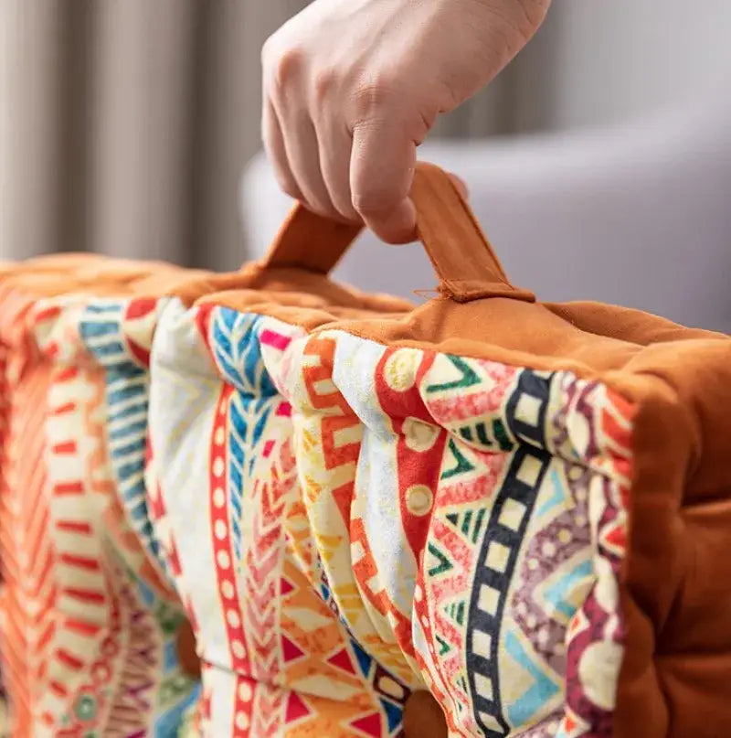 A cozy bohemian mandala velvet floor cushion with vibrant quilted geometric orange red blue cream pattern and brown handle