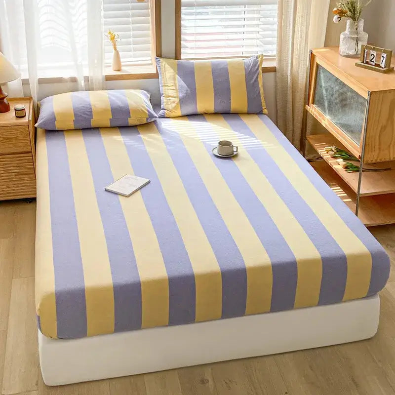 A cozy bed with a lavendar and yellow striped decorbites cotton stripe fitted sheet and matching pillowcases