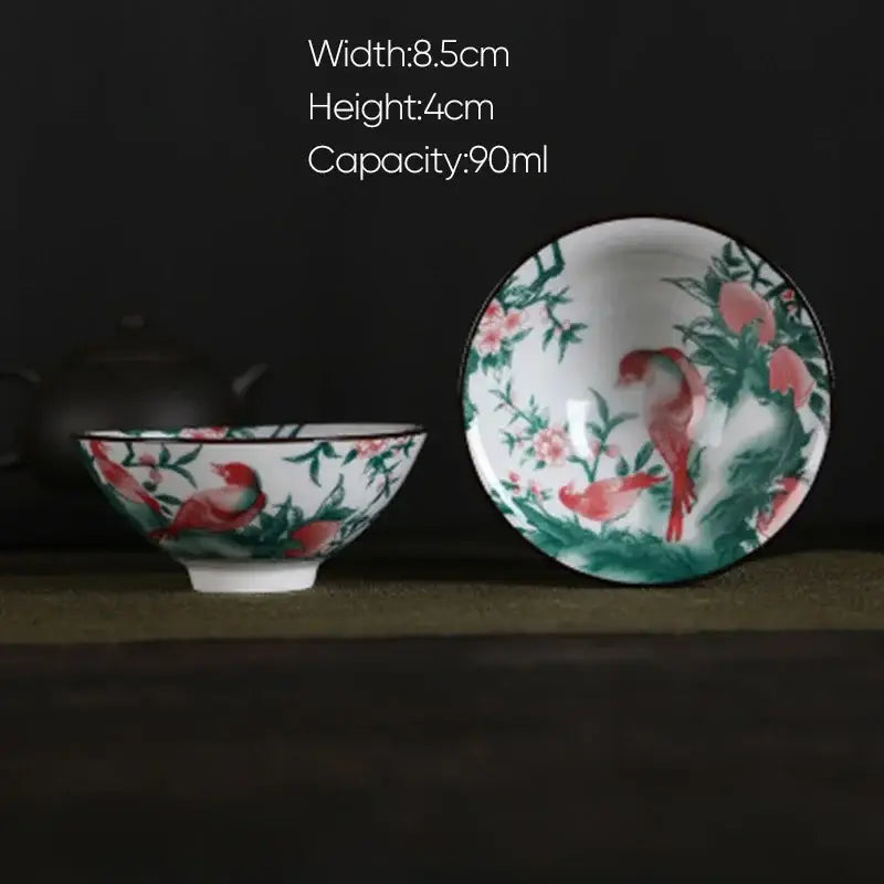 A delicate white porcelain hand-painted tea bowl with vibrant red birds and green foliage in a pink and black floral set