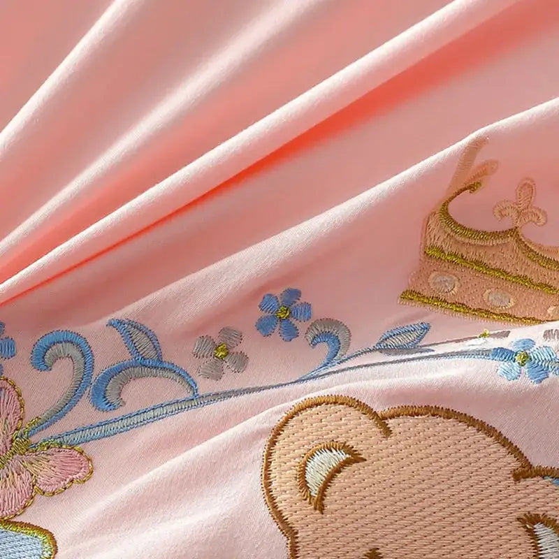 Pink satin embroidered bed skirt set featuring blue gold and beige floral and crown motifs for bedroom decor