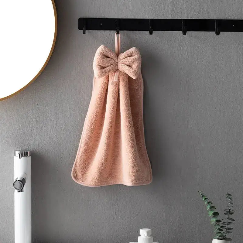 A soft peach coral velvet hand towel with a plush bow hanging on a black hook for kitchen and bathroom decorbites